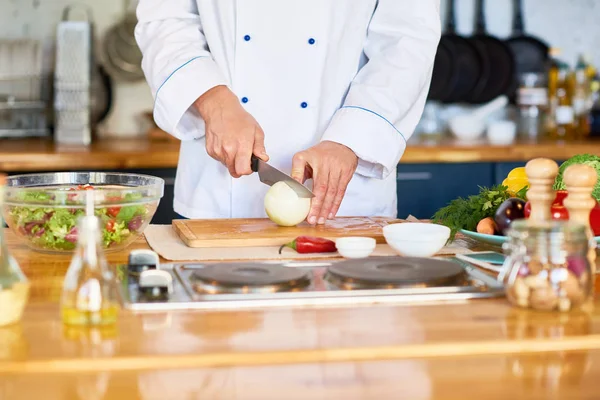在准备素食沙拉时 穿着制服的男厨师站在餐厅厨房和切洋葱的餐桌上 — 图库照片