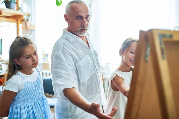 Portrait of mature art teacher teaching children painting in art class explaining techniques and pointing at easel