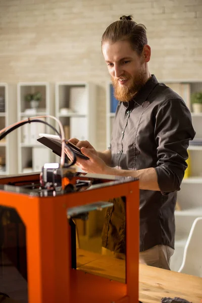 Portrait of handsome young man enjoying work with 3D printer in modern design studio, using digital tablet and smiling