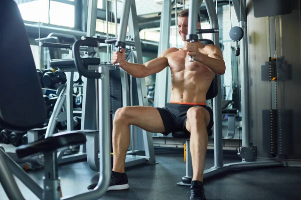 Full length portrait of handsome muscular man with bare chest exercising on machine during workout in modern gym