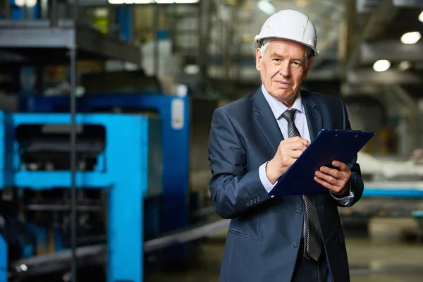 Portrait of senior businessman wearing hardhat looking at camera holding clipboard in workshop of modern factory