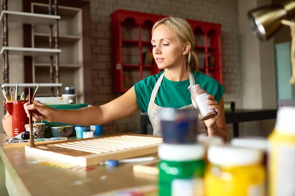 Portrait of pretty blonde woman enjoying work in art studio painting shutters with bronze paint, making DIY interior decoration