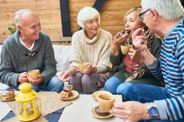 Group of elderly men and women warming themselves with hot tea and enjoying delicious cakes while having gathering at cozy small patio