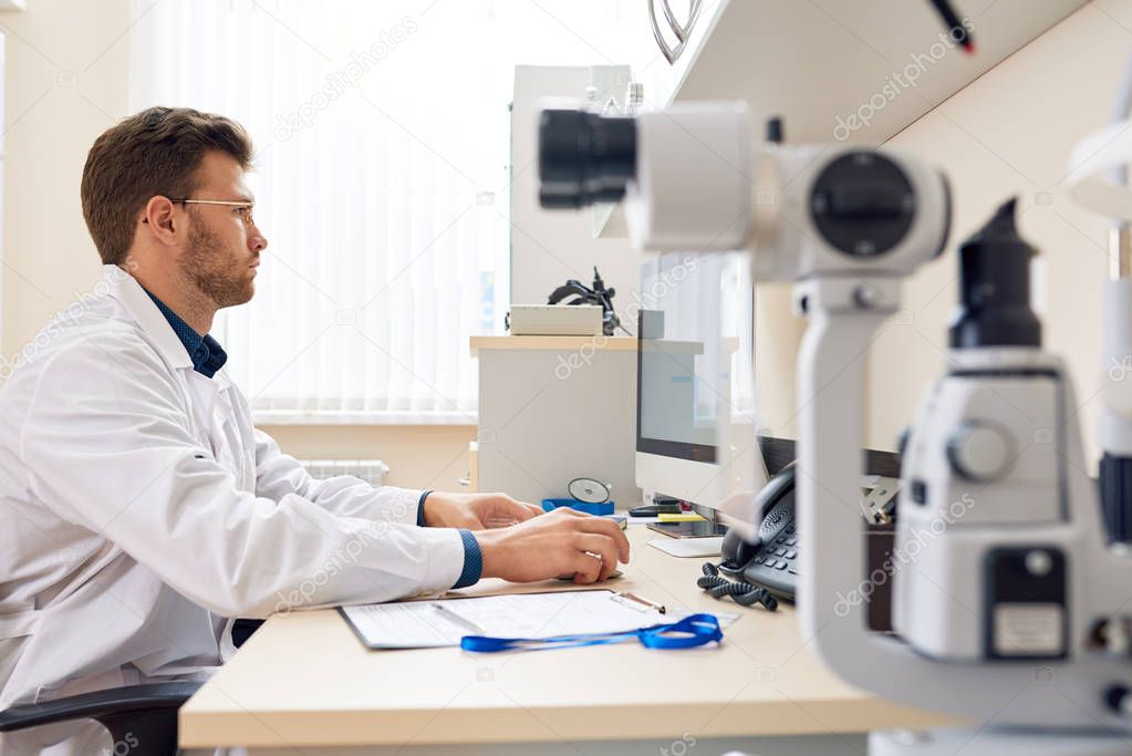 Side view portrait of  male ophthalmologist  using PC sitting at desk in office and typing on keyboard, optometric equipment in foreground