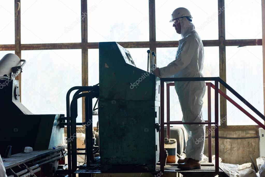Side view full length of worker wearing biohazard suit operating massive machine in factory workshop standing against window