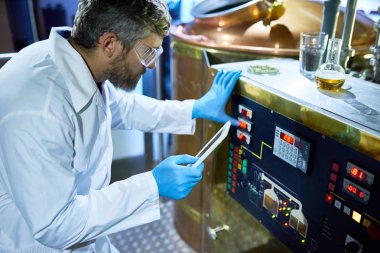 Ayar makine yardımıyla dijital tablet, iç arka plan üzerinde modern bira fabrikası, ekranda sunulan kullanım kılavuzu, orta yaşlı brewer Beyaz önlük ve koruyucu gözlükler