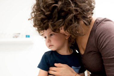 Kıvırcık annesi kucağa oturma ve uzakta o onu yanak, beyaz arka plan üzerinde öpüşme süre seyir tatlı küçük çocuk portre vurdu