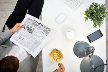 Esmer girişimci öğle yemeğinde Ofis Masası zevk: o gazete okuma ve yemek çubukları, hemen üstündeki Görünüm ile anlık erişte yemek