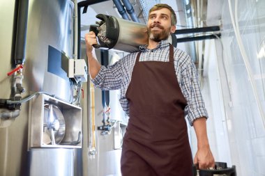 Gülümseyen portre çekim modern Fabrikası'nın koridordan yürürken ağır bira fıçıları taşıyan önlük giyen brewer sakallı