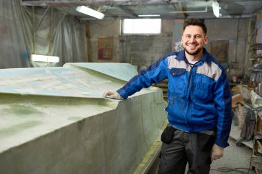 Portrait of modern mature man smiling happily at camera while posing in yacht workshop next to repaired boat clipart