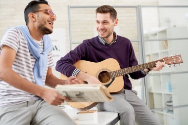 İki kaygısız genç adam gitar ve masada oturan şarkılar çalmaya Office eğleniyor portresi
