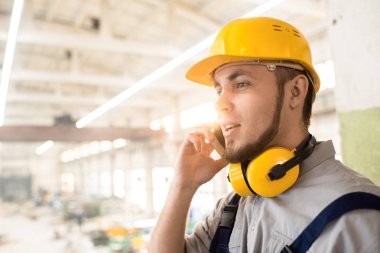 Modern bitki geniş üretim departmanı ayakta ve onun üstün mobil telefonda konuşurken hardhat ve kulak koruyucular giymiş yakışıklı sakallı işçi baş ve omuzlar portresi.