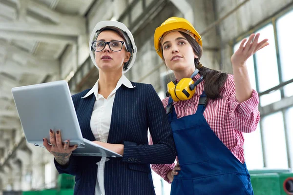 指すとラップトップを保持している近代的な工場の広々 としたワーク ショップに立って Hardhats を身に着けている 人の女性エンジニアの肖像画 — ストック写真