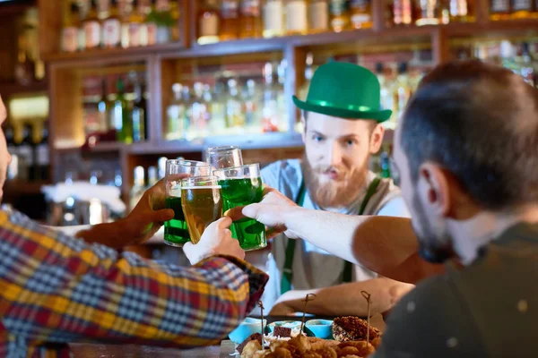 欢快的年轻人在现代酒吧闲逛时用啤酒杯举杯 英俊的大胡子酒保身穿绿色圆顶帽子 — 图库照片
