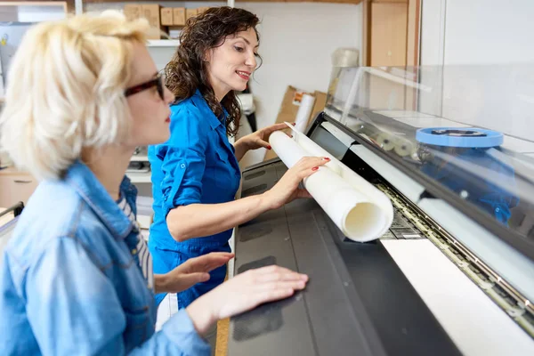 Side view portrait of two smiling young women working with plotter machine in printing shop or publishing company