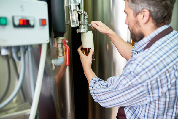 Concentrated brewing engineer in shirt pouring beer into glass while examining quality of producing product at brewery factory