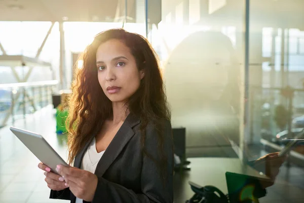 Confident mixed race entrepreneur in formalwear looking at camera while standing at spacious office lobby with panoramic windows and using digital tablet, waist-up portrait