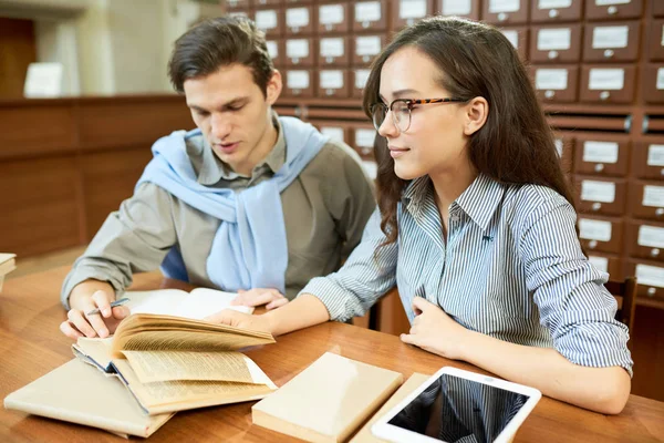 Group of hard-working students writing report together while sitting at wooden desk of reading room, catalogue cabinets on background