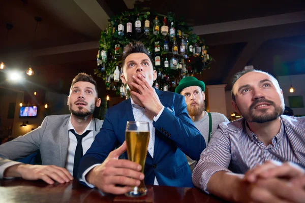 一群集中的朋友看足球比赛 而坐在酒吧柜台和喝啤酒 胡子男子穿着妖精服装站在他们身后 — 图库照片