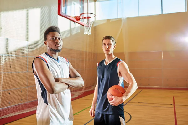 Serious confident professional basketball players ready for game: handsome African-American guy with beard crossing arms, Caucasian man holding ball