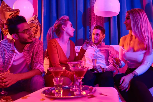 Group of people chilling on sofa during luxurious house party, drinking alcoholic beverages and chatting in room lit by dim blue light