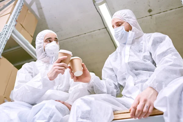 男性仓库工人穿着安全口罩和工作服的低角度视图在喝咖啡的时候 — 图库照片