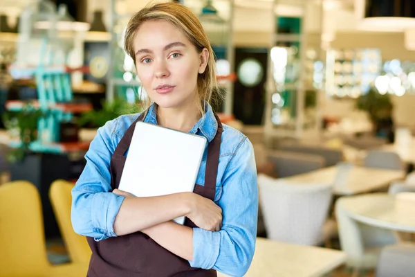Waist-up portrait of pretty blond-haired waitress wearing apron posing for photography while holding digital tablet in hands, interior of spacious restaurant on background