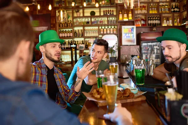 一群英俊的年轻朋友们在现代酒馆里享受着彼此的陪伴 他们喝啤酒 吃零食和聊天 其中一个和胡子酒保戴着绿色的帽子 — 图库照片