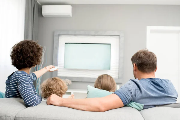 リビング ルームのソファーに座っていると 現代のテレビの電源を入れる設定リモート コント ローラーとテレビを見ている 人の子供と幸せな家族の肖像をビューをバックアップします — ストック写真