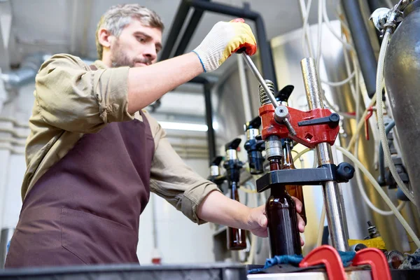 工場直売店でビール瓶を閉じながらキャップ締めを使用して手袋で作業エプロンで忙しい熟練した残忍な醸造技師 — ストック写真