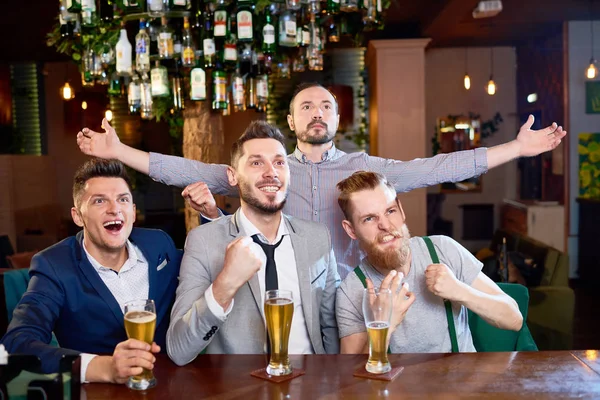 Joyful group of friends celebrating victory of their favorite football team while sitting in modern pub and drinking beer