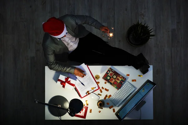 仕事でクリスマス イヴを祝う 認識できないホワイト カラー労働者の足を机の上に座っていると近代的な薄暗いオフィスのインテリアを手に 花火線香花火を保持のサンタ帽子をかぶって — ストック写真