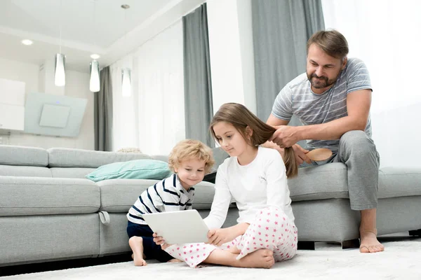 Lovely family of three gathered together in cozy living room and watching their favorite TV show on digital tablet, bearded middle-aged father distracted from combing hair of his adorable daughter