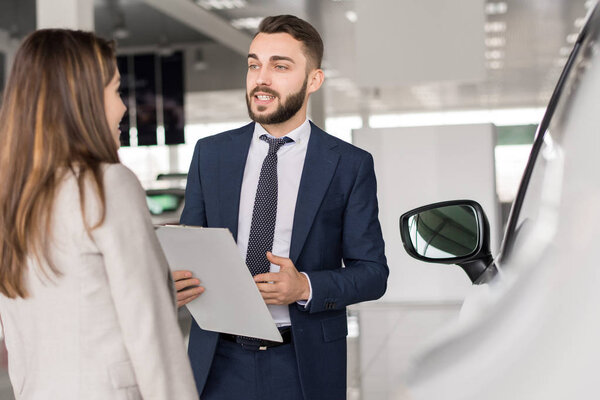 Portrait of handsome car salesman talking to young woman helping her choose in car showroom