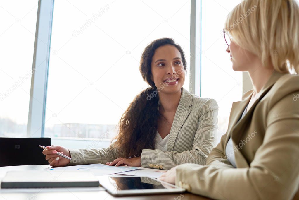 Pretty mixed race manager with long curly hair and her blonde colleague gathered together in office lobby and sharing creative ideas with each other