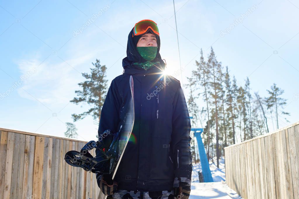 Low angle portrait of modern young man holding snowboard and looking away with determination standing against clear blue sky, lens flare