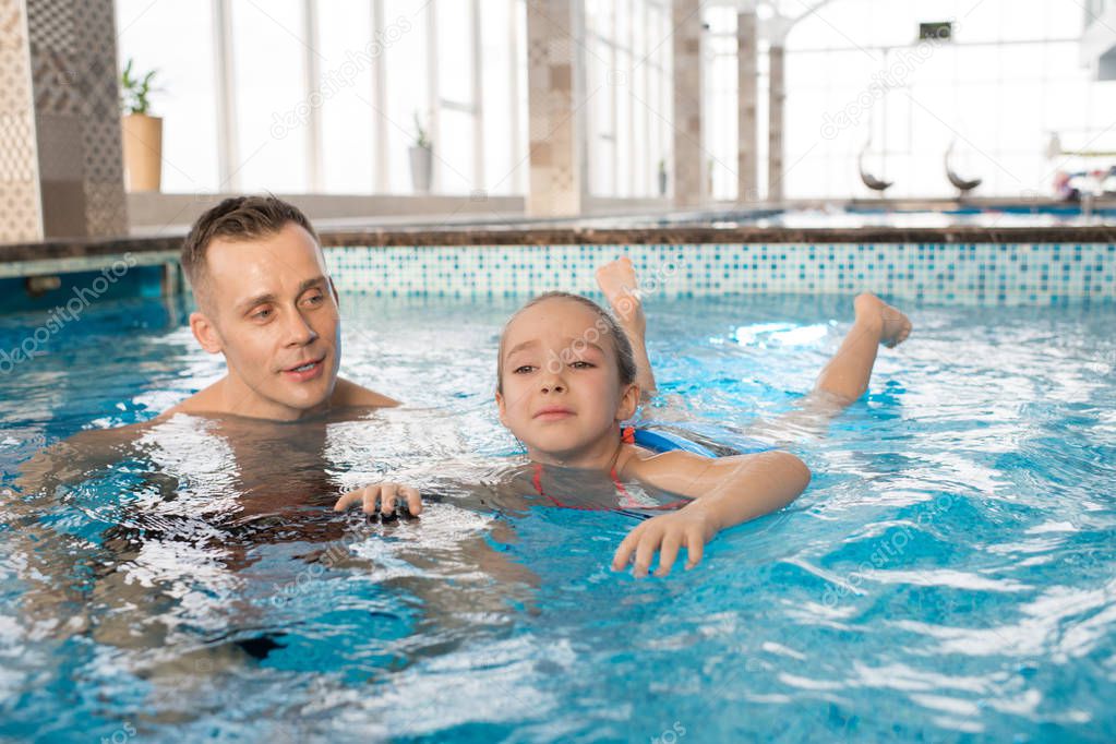 Portrait shot of handsome man teaching his frightened little daughter to swim in modern swimming pool with panoramic windows
