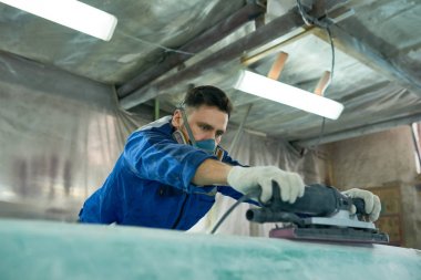 Portrait of mature modern man wearing respirator repairing boats in workshop using electric polishing tool clipart