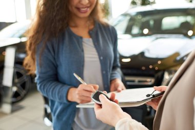 Close-up shot of smiling curly woman signing contract for new car, unrecognizable saleswoman holding key in hand, interior of modern showroom on background clipart