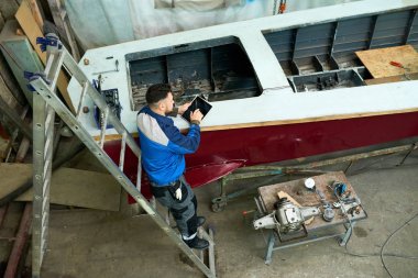 High angle portrait of modern worker using digital tablet while repairing boat in yacht workshop clipart