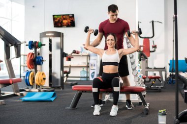 Kişisel Spor hocası tarafından yardım bankta oturmuş dumbbells ile egzersiz güzel genç kadın portresi tam uzunlukta