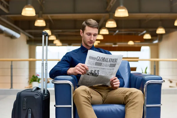 Portrait of handsome young man with suitcase reading newspaper sitting in leather armchair while waiting for check in in modem hotel