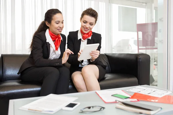 两个穿着黑色和红色制服的开朗女人的肖像使用数字片和微笑 而坐在沙发上 在现代办公室和看设备屏幕 — 图库照片