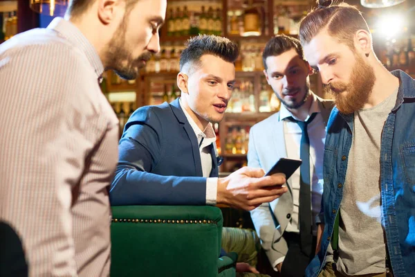 Group of bearded friends gathered together in pub after hard working day, chatting with each other and looking through photos on smartphone