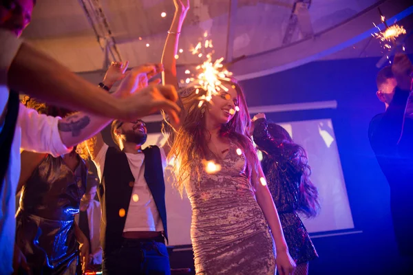 一群时髦的年轻人在夜总会里庆祝节日在舞池中间燃烧着熊熊的烟火 专注于跳舞的漂亮女孩穿着闪闪发亮的天鹅绒派对礼服 — 图库照片