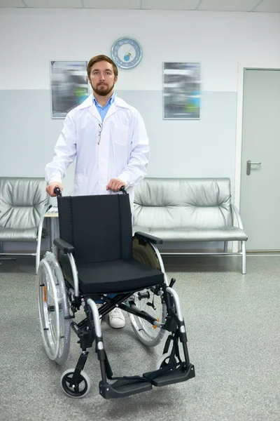 Portrait of young bearded doctor standing in hospital hall posing with empty wheelchair and looking at camera