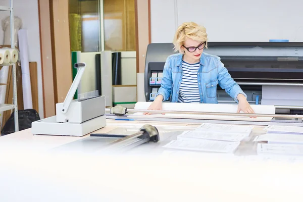 Portrait of blonde young woman working in modern printing shop or publishing company, cutting  paper and loading plotter machines