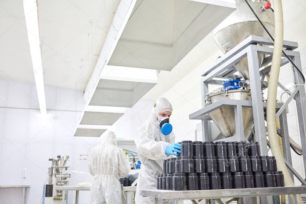 Two pharmaceutical factory workers wearing coveralls and respirators wrapped up in preparations for product shipment, interior of manufacturing department on background