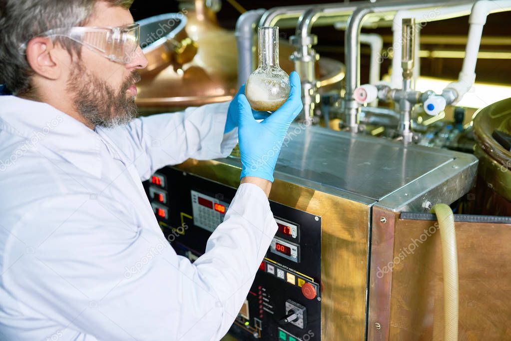 Profile view of confident middle-aged brewer wearing white coat, rubber gloves and protective goggles holding flask with beer in hands while checking its quality