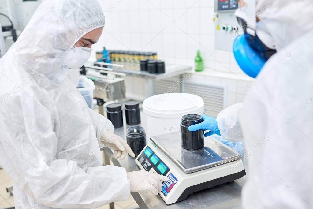 Profile view of concentrated factory worker wearing overall and safety mask using electronic scale while filling in plastic bottle with sport nutrition